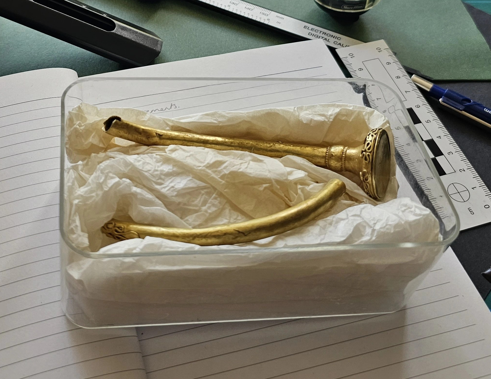 A gold broken torc in a plastic box, resting in tissue paper. The box is sat on a notebook