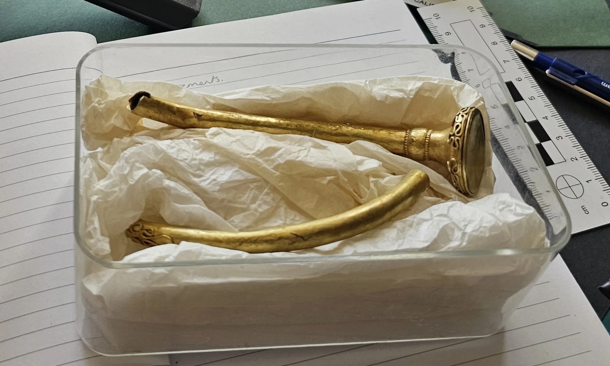 A gold broken torc in a plastic box, resting in tissue paper. The box is sat on a notebook